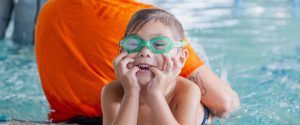 Child in green goggles leans on the side of the pool