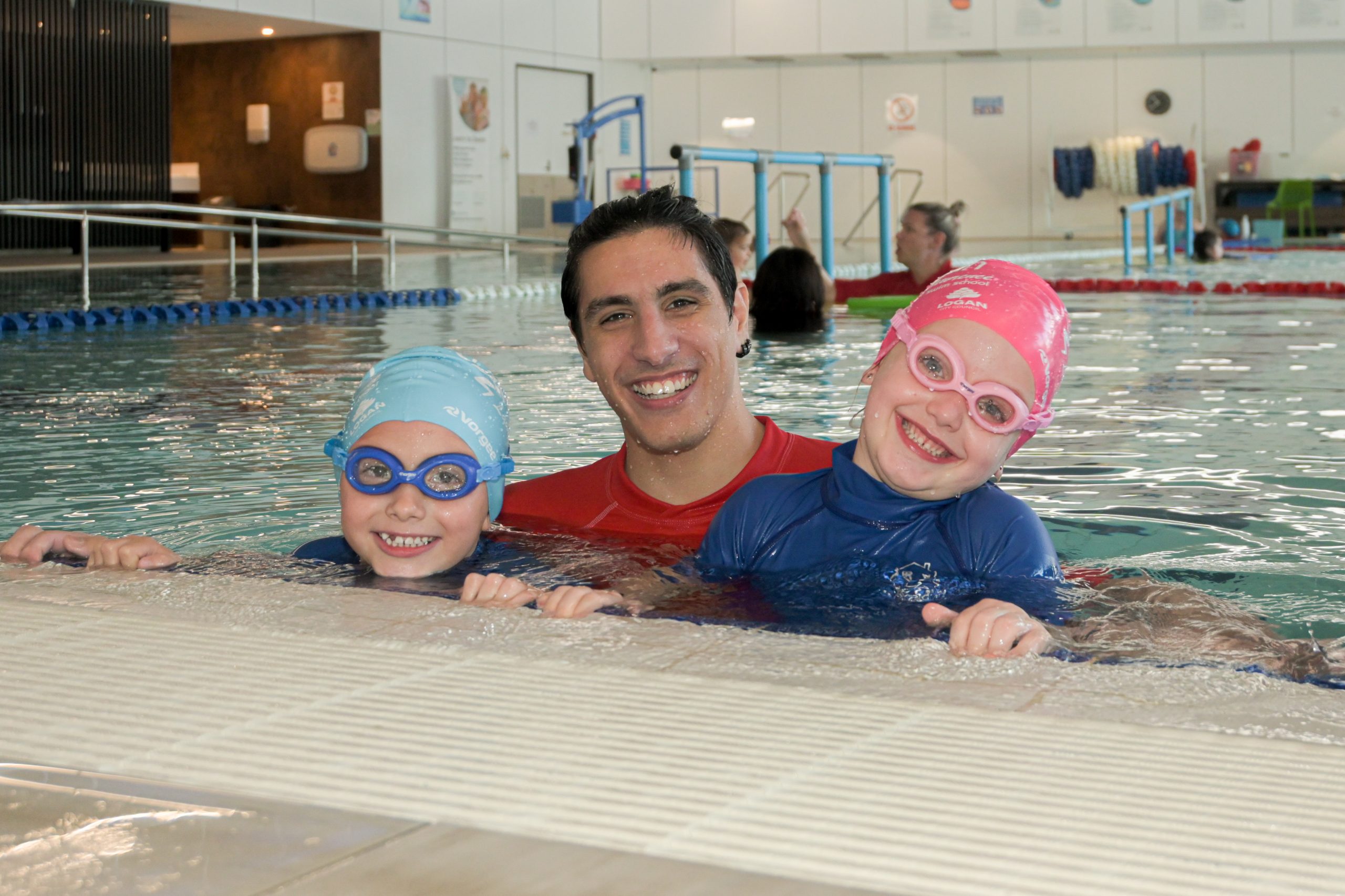 Logan Swim School Tutor smiles with a student either side of him leaning on the edhe of the indoor pool at Beenleigh Aquatic Centre