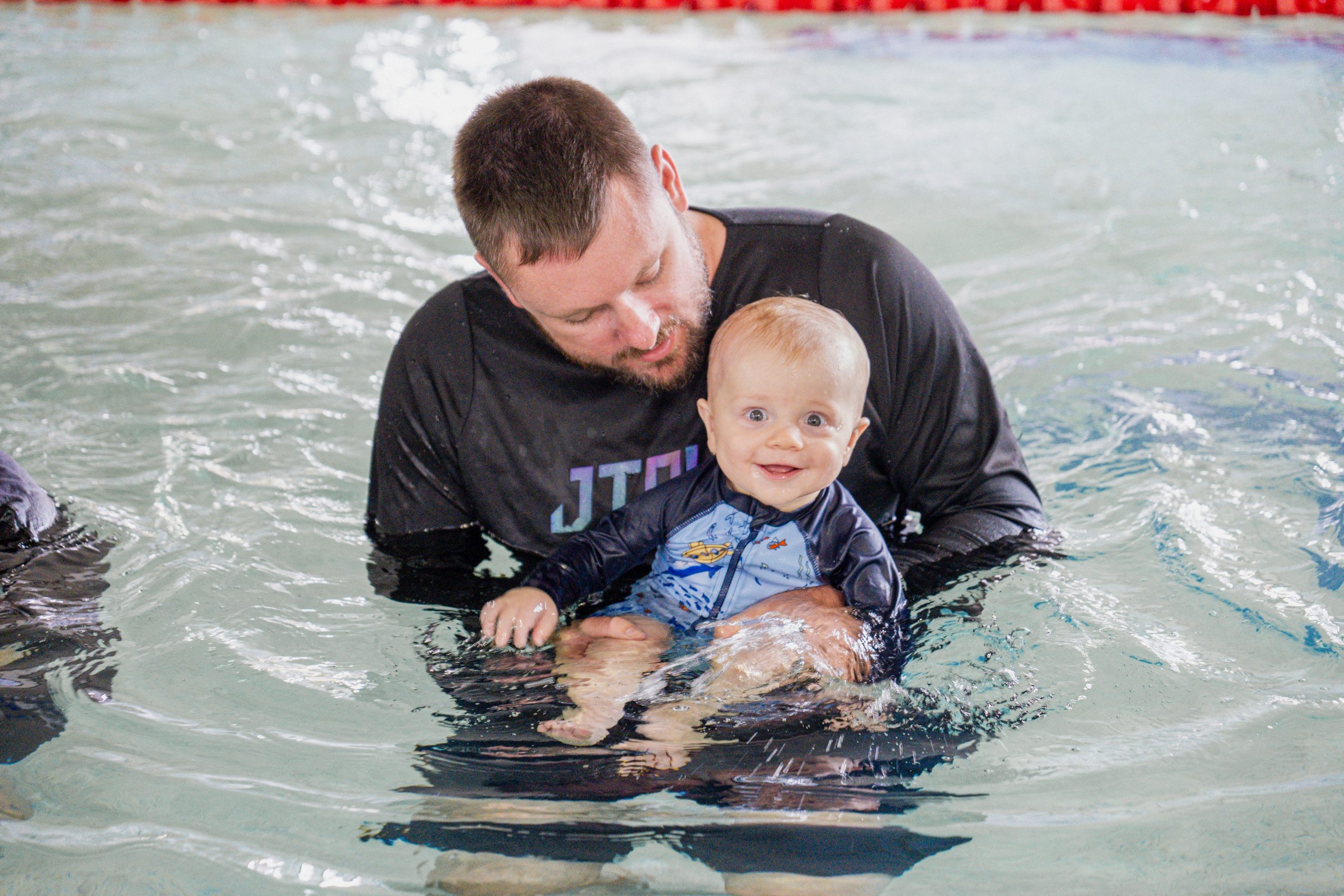 Young boy baby smiles at camera while Dad in black swim shirt holds him in the water