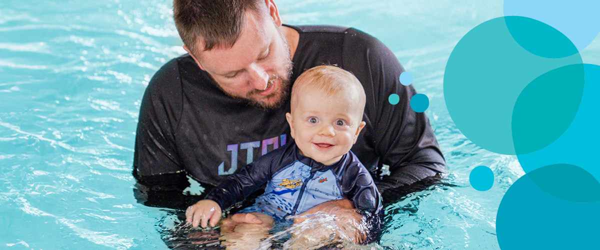 Young boy baby smiles at camera while Dad in black swim shirt holds him in the water